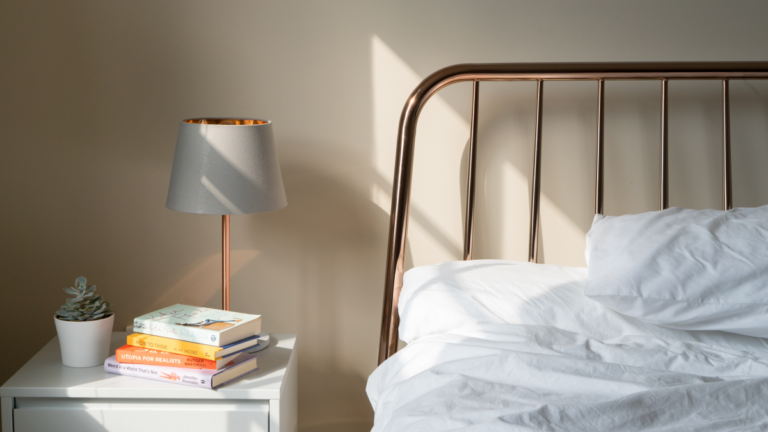 Imagem da cabeceira de uma cama com uma mesa de apoio ao lado e um abajur em cima.