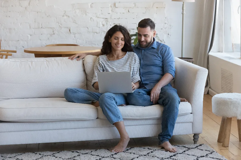 Um casal sentado confortavelmente no sofá enquanto a mulher segura um notebook, pesquisando informações sobre "como comprar um imóvel em casa"